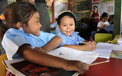 Niños en uniforme azúl sentados en un salón de clase con cuadernos en frente de ellos.