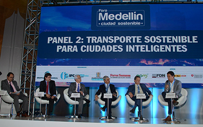 Seis hombres en traje sentados en sillas blancas en un escenario. La pared dice, “Foro Medellín ciudad sostenible” y “Panel 2: Transporte sostenible para ciudades inteligentes”.