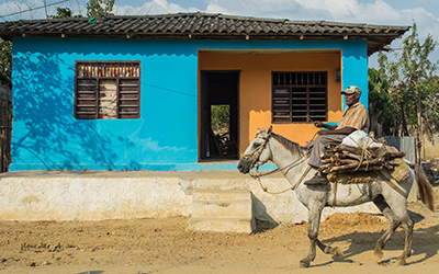 Un hombre montado a caballo en un camino de tierra y una casa azul detrás de él.