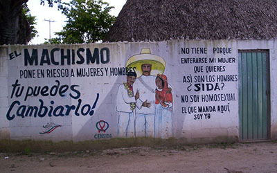 Un mural en una pared blanca que dice, “el machismo pone en riesgo a mujeres y hombres tu puedes cambiarlo” a la izquierda y “no tiene porque enterarse mi mujer, que quieres, así son los hombres, ¿sida?, no soy homosexual, el que manda aquí soy yo” a la derecha. En el centro hay tres figuras con vestimenta campesina tradicional.