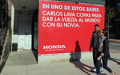 Un hombre y una mujer caminan en la acera por un cartel rojo en la pared que dice “En uno de estos bares Carlos lava copas para dar la vuelta al mundo con su novia”. En la parte baja del cartel dice, “Honda The Power of Dreams”.
