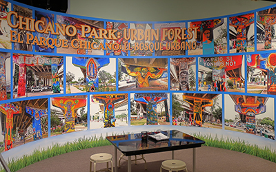 En el fondo hay imágenes del Parque Chicano con texto que dice “Chicano Park Urban Forest, Parque Chicano Bosque Urbano”. En el centro hay una mesa negra con tres banquillos blancos.