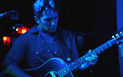 Gustavo Galindo tocando la guitarra en un escenario azul oscuro con un micrófono al lado.