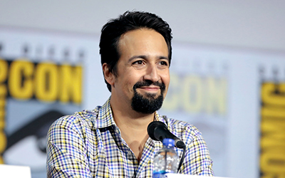 Lin Manuel Miranda con una camisa cuadriculada sentado en una mesa con un micrófono para el evento de Comic-Con.
