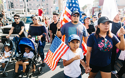 Hispanos marchan en la calle. El hombre y la mujer a la izquierda empujan coches con un niño y una niña y las personas a la derecha tienen banderas estadounidenses en las manos.