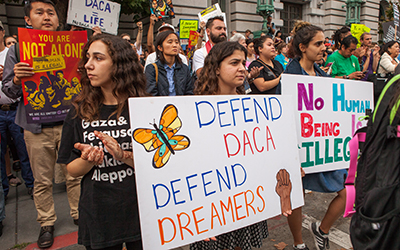 Personas en la calle en San Francisco en una manifestación para DACA con pancartas que dicen, “Defend DACA Defend Dreamers” y “No Human Being is Illegal”.