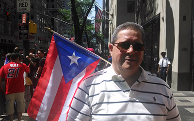 Un hombre en la calle con una bandera de Puerto Rico y con un policía y otras personas al fondo.