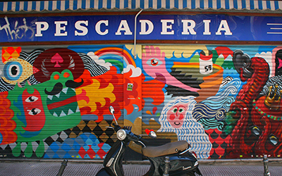 Graffiti de diferentes animales de colores vibrantes en una tienda que dice “pescadería”. Hay una moto estacionada en la calle enfrente del mural.