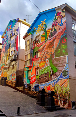 Al lado derecho de una calle pedestre en Vitoria-Gasteiz, hay dos edificios cuyas paredes están completamente cubiertas por murales muy coloridos. Los murales contienen una mezcla de estilos artísticos diferentes.