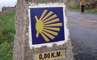 Una piedra alta al lado de la calle que tiene la concha de vieira en amarillo en un trasfondo azul, y escrito por debajo “0,00 K.M.”.
