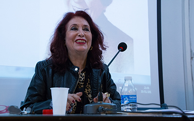 Lidia Falcón sonriendo sentada en una mesa con un micrófono, un vaso blanco de plástico y una botella de agua. En el trasfondo hay una pantalla de proyección.