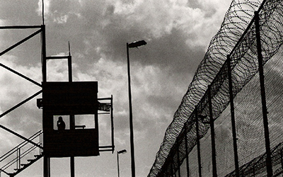 Una valla con alambre de púas y una persona en una torre de vigilancia todo en blanco y negro.