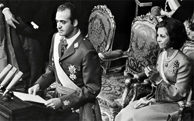 Una imagen en blanco y negro de el Rey Juan Carlos ante un podio y Sofía sentada atrás de él.