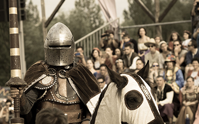 Un caballero sosteniendo una lanza montado en un caballo con espectadores sentados en bancas a sus espaldas.