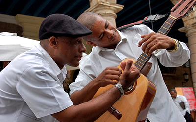 Dos hombres vestidos de blanco observando y tocando la misma guitarra.
