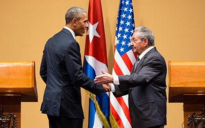 El presidente Obama y el presidente Castro dándose un apretón de manos con la bandera cubana y estadounidense en el fondo.