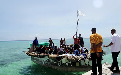 Una balsa llena de inmigrantes en el mar con un hombre alzando un pañuelo blanco y dos hombres parados en el muelle.