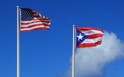 La bandera estadounidense cuelga del asta a la par de la  bandera puertorriqueña colgada del asta bajo un cielo azul.