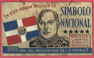 Un cuadro de madera con la bandera de la República Dominicana y la imagen de Trujillo y que dice, “En este hogar Trujillo es símbolo nacional” en azul  y “rectitud, libertad, trabajo y moralidad” en rojo, y “1955 año del benefactor de la patria” en azul.