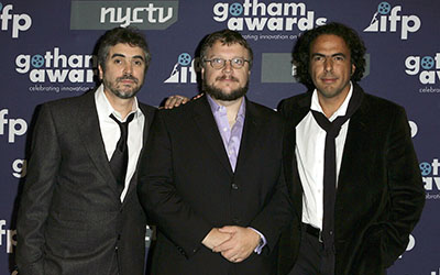 Alfonso Cuarón, Alejandro Iñárritu y Guillermo del Toro con trajes negros en frente de una pared que dice “gotham awards”.