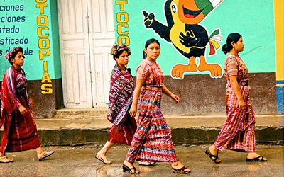 Cuatro mujeres con vestimenta tradicional guatemalteca caminando en frente de un edificio con una pared verde que dice, “fotocopias” y que tiene un dibujo de un pájaro.