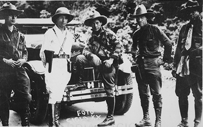 Una imagen en blanco y negro del general Sandino y otros hombres en uniforme parados en frente de un coche.