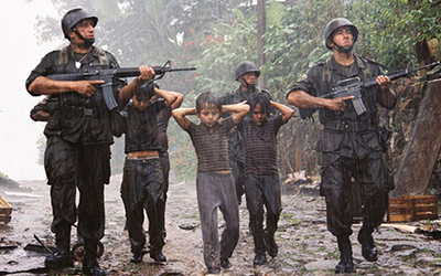 Tres soldados con armas acompañan a tres niños con las manos en la cabeza en un día lluvioso.