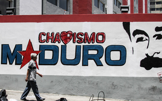 Un mural en la pared que dice “Chavismo Maduro”, pero la “v” ha sido reemplazada por un corazón, y la “a” por una estrella. El perfil de Maduro está a la derecha y un hombre camina enfrente del mural.