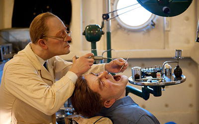 Un dentista con una bata blanca sentado en una silla y un paciente con una camisa azul con la boca abierta mientras el dentista le examina la boca.