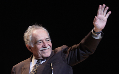 Gabriel García Márquez con un saco negro sonriendo y saludando con su mano izquierda.
