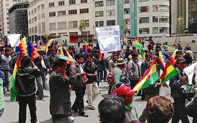 Personas marchando en la calle con pancartas y la bandera wiphala.