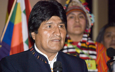 Evo Morales en una chaqueta y un indígena y una bandera al fondo.