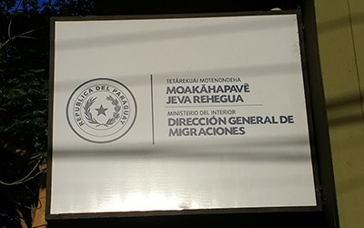 Un cuadro con orilla negra con texto en guaraní/español en el centro que dice “Dirección general de migraciones”.