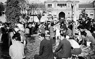  Una fotografia en blanco y negro de personas (simpatizantes de Juan Perón) sentadas a la orilla de la fuente en la Plaza de Mayo.