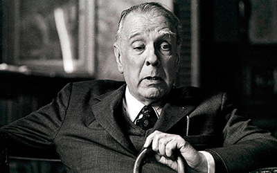 Una imagen en blanco y negro de Jorge Luis Borges con un saco sentado en su oficina con un bastón en la mano.